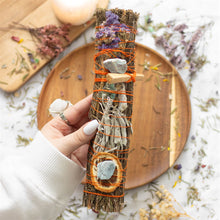 Load image into Gallery viewer, A person holding a smudge wand over a wooden plate. The wand is hand tied with dried orange slices, rosemary and adventurine stone. The wooden plate is surrounded by sprigs of dried herbs and flowers.