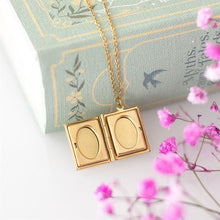 Load image into Gallery viewer, A gold-toned book-shaped pendant necklace. The locket is opened to show the oval windows used to display photos. The gold toned chain hangs over a blue decorative book with pink flowers in the foreground.