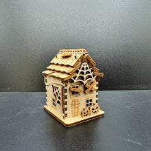Load image into Gallery viewer, A laser cut haunted house model, side on shot. With boarded up windows and a large cobweb across the peak of the roof. Two tiny pumpkins decorate the front of the house. The front door is shaped like a coffin. The side wall reveals a hinge so the house can open up to store small items inside. The roof is designed like layered tiles. Tiny wooden boards cover any gaps where the tiles mimic dilapidated fallen tiles.