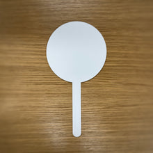Load image into Gallery viewer, A single white laser cut acrylic sign with a handle, it is circular shaped, placed on a wooden surface.