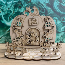 Load image into Gallery viewer, A wooden laser cut fairy door shaped like a pumpkin with layered vine leaf detail, four small windows, a layered hinge on the door (This version is only decorative and is not a working door), and a small engraved sign with the words "Trick or Treat" above the door. The item free-stands on a mock stone floor base with vine styled fencing in front of it.