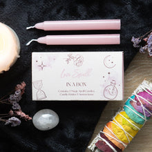 Load image into Gallery viewer, A Spell in a box gift set to promote Love on a black background with two of the candles that come in product. The background is decorated with sage sticks, crystals, flowers and other candles.