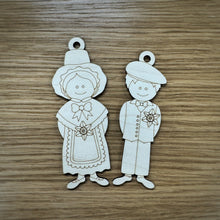 Load image into Gallery viewer, Two laser cut wooden tags of a people in traditional Welsh outfits. Each have a loop on top of their heads. Items on a wooden surface.