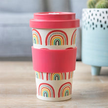 Load image into Gallery viewer, A reusable travel tumbler with a cream-coloured body printed with colourful rainbow patterns, a bright red lid and a matching red silicone grip band in the middle. Item on a light wooden surface with a blue spotted plant pot and a sofa seen in the background.