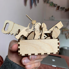 Load image into Gallery viewer, A hand holding a laser cut wooden desktop hobby set consisting of various wooden tool shapes such as a saw, wrench, a hammer, a drill, and others inside a a small wooden tool box.