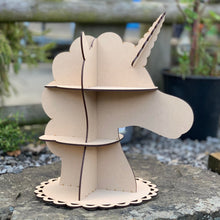 Load image into Gallery viewer, An MDF laser cut unicorn bust cupcake stand. The stand has a side and front profile silhouette that slots together. The circular base has a scalloped rim. Item photographed on a stone wall in a garden.