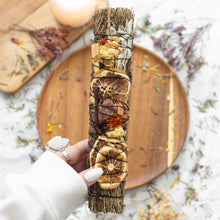 Load image into Gallery viewer, A person holding a smudge wand over a wooden plate. The wand is hand tied with dried orange slices, rosemary and red jasper stone. The wooden plate is surrounded by sprigs of dried herbs and flowers. 

