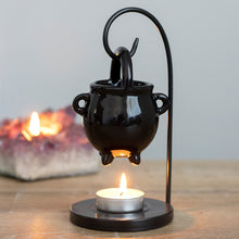 Load image into Gallery viewer, A Ceramic black cauldron-shaped tealight oil/wax burner hanging on a metal stand with a tea light lit on the base. Item is displayed on a wooden table with a lit candle in the background.
