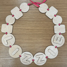 Load image into Gallery viewer, A wooden laser cut friendship bracelet bunting with a pink ribbon through the varying bead shapes and the name "LIZZIE" layered on the circular beads. Items on a darker wooden background.
