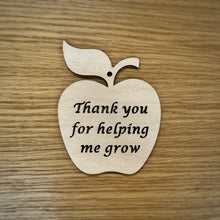 Load image into Gallery viewer, A laser cut wooden apple shape with cut out text reading "Thank you for helping me grow" and hole cut out at the top for hanging. Item resting on a darker wood grain surface.
