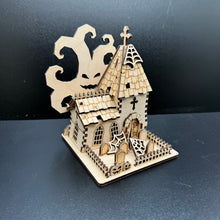 Load image into Gallery viewer, Laser cut halloween themed church model. A spooky tree with a pumpkin style face behind the building. In front is a graveyard garden surrounded by a jagged fence. Cobwebs are scattered in the garden and on the church spire.
