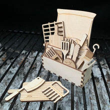 Load image into Gallery viewer, A laser cut set of BBQ tool &amp; cutlery shapes inside a wooden tool box. The set is displayed on a real BBQ. 
