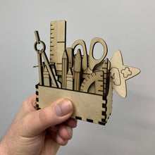 Load image into Gallery viewer, A hand holding a tool box filled with laser cut stationery shapes, such as a ruler, a protractor, a pair of scissors, pens, pencils and other items.
