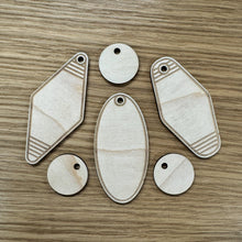Load image into Gallery viewer, Six laser cut keyring shapes. Three small circular keyrings to emulate the round room number tag on hotel keys, with three different shaped keyrings in vintage styles. One in a diamond shape, another in an oval shape, and the last in a classic motel shape. (Like a diamond but with round corners)
Twp of the larger motel keyrings have an engraved border with several lines at the top and bottom, the oval shaped only has a border. Items displayed on a wooden background.
