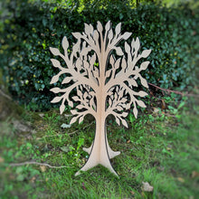Load image into Gallery viewer, A large laser-cut MDF free standing tree made from two flat pieces that interlock together, upright on a grassy lawn with a blurred green hedge background.
