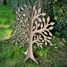 Load image into Gallery viewer, A large laser-cut MDF free standing tree made from two flat pieces that interlock together, upright on a grassy lawn with a blurred green hedge background.
