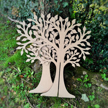 Load image into Gallery viewer, Two laser cut MDF tree shapes, leaning against a real tree in a garden to show the two parts of the interlocking free standing tree.
