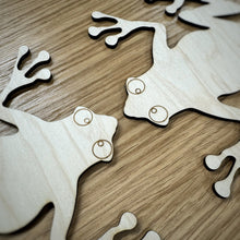 Load image into Gallery viewer, A close up of the heads of two wooden laser cut frog shapes on a wood surface.
