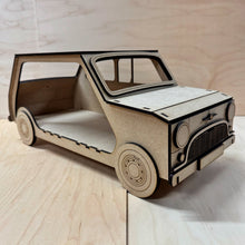 Load image into Gallery viewer, The front corner of a 3D MDF laser cut cupcake stand in the shape of a classic mini car. Item has layered lights, bumpers, grill, logo and other parts and this right side is open to allow extra storage for more cupcakes. Item on a light wooden background.
