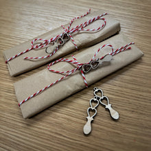 Load image into Gallery viewer, Two small craft paper wrapped parcels with red and white jute string tied in a bow around both. Each have a mini laser cut love spoon tied to the string. Two mini spoons are laid out in front of the parcels. 
Everything is on a wooden surface.
