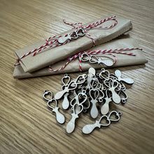 Load image into Gallery viewer, Two small craft paper wrapped parcels stacked on top of each other with red and white jute string tied in a bow around both. Each have a mini laser cut love spoon tied to the string. The rest of the eighteen mini spoons are laid out in front of the parcels. 
Everything is on a wooden surface.
