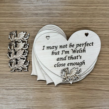 Load image into Gallery viewer, A stack heart shaped laser cut plaques, featuring two heart shaped holes at the top for hanging, with the following phrase cut out: "I may not be perfect, but I'm Welsh and that's close enough". A laser cut Welsh Dragon is cut out and layered at the bottom of the heart plaque at the top of the stack. Another four dragon shapes are positioned next to the plaques to show they're a part of the set.
