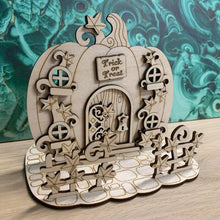 Load image into Gallery viewer, A wooden laser cut fairy door shaped like a pumpkin with layered vine leaf detail, four small windows, a layered hinge on the door (This version is only decorative and is not a working door), and a small engraved sign with the words "Trick or Treat" above the door. The item free-stands on a mock stone floor base with vine styled fencing in front of it.
