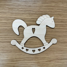 Load image into Gallery viewer, A wooden laser cut rocking horse shape. The design has a cut out heart on the body and smaller hearts on the base. Item on a wooden background.
