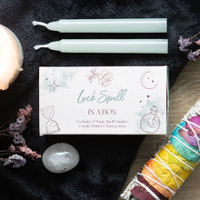 Load image into Gallery viewer, A Spell in a box gift set to promote Luck on a black background with two of the candles that come in product. The background is decorated with sage sticks, crystals, flowers and other candles.
