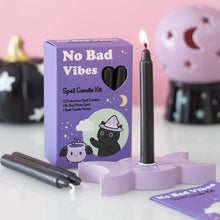 Load image into Gallery viewer, A purple box with a black cat cartoon over a cartoon cauldron with bat wings on the cover. At the top: "No Bad Vibes" is written in white and below that: "Spell Candle Kit" Along with the contents of the box in black text: "12 Protection Spell candles. 1 no bad vibes spell and 1 spell candle holder." A candle is lit and displayed inside the triple moon of two crescents and one full moon candle holder, which has a pentagram on the centre. Two more candles are resting next to the box. 
