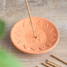 Load image into Gallery viewer, A terracotta incense stick holder plate with an embossed sun and face design, holding a plain incense stick.
