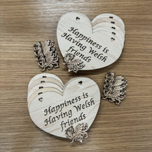 Load image into Gallery viewer, Two stacks of heart shaped laser cut plaques with a layered Welsh dragon shape on top of each stack. The plaque design has two heart shaped holes cut out at the top for hanging and the following phrase cut out: "Happiness is Having Welsh friends". Next to the stacks are more dragons to layer on the other plaques. Items on a wooden background.
