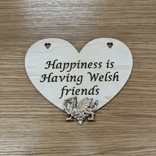 Load image into Gallery viewer, A heart shaped laser cut plaque with a layered Welsh dragon shape on top. The plaque has two heart shaped holes cut out at the top for hanging and the following phrase cut out: "Happiness is Having Welsh friends". Items placed on a wooden background.
