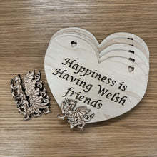 Load image into Gallery viewer, A stack of heart shaped laser cut plaques with a layered Welsh dragon shape on top. The plaque has two heart shaped holes cut out at the top for hanging and the following phrase cut out: "Happiness is Having Welsh friends". Next to the stack are more dragons to layer on the other plaques. Items on a wooden background.
