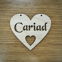 Load image into Gallery viewer, A heart-shaped wooden sign with the word 'Cariad' cut out in a Celtic stencil font and a smaller heart cut out below. Two holes at the top of the heart are cut out for hanging the heart. Item displayed on a wooden surface.
