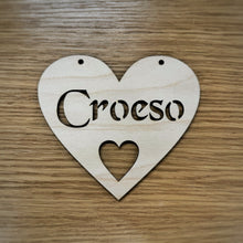 Load image into Gallery viewer, A heart-shaped wooden sign with the word 'Cariad' cut out in a Celtic stencil font and a smaller heart cut out below. Two holes at the top of the heart are cut out for hanging the heart. Item displayed on a wooden surface.
