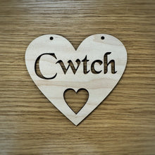 Load image into Gallery viewer, A heart-shaped wooden sign with the word 'Cwtch' cut out in a Celtic stencil font and a smaller heart cut out below. Two holes at the top of the heart are cut out for hanging the heart. Item displayed on a wooden surface.

