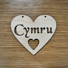 Load image into Gallery viewer, A heart-shaped wooden sign with the word 'Cymru' cut out in a Celtic stencil font and a smaller heart cut out below. Two holes at the top of the heart are cut out for hanging the heart. Item displayed on a wooden surface.
