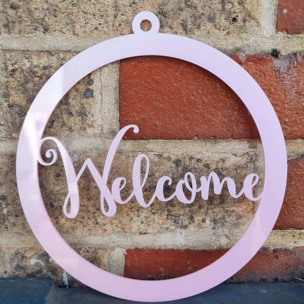A laser cut pink-acrylic sign with the word welcome in the center, with a brick wall in the background.