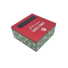 Load image into Gallery viewer, A Christmas themed box for pets. It features a green box with a white paw print pattern and a red lid. On the top of the box there is a black chalkboard panel to write in your pet's name and below that is a white paw and the words "Believes in Santa Paws". There's a gold clasp on the front and the lid is hinged. Item on a white background.
