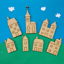 Load image into Gallery viewer, A collection of laser cut toy box village building shapes placed against a blue &amp; green background like a hill with white clouds.
